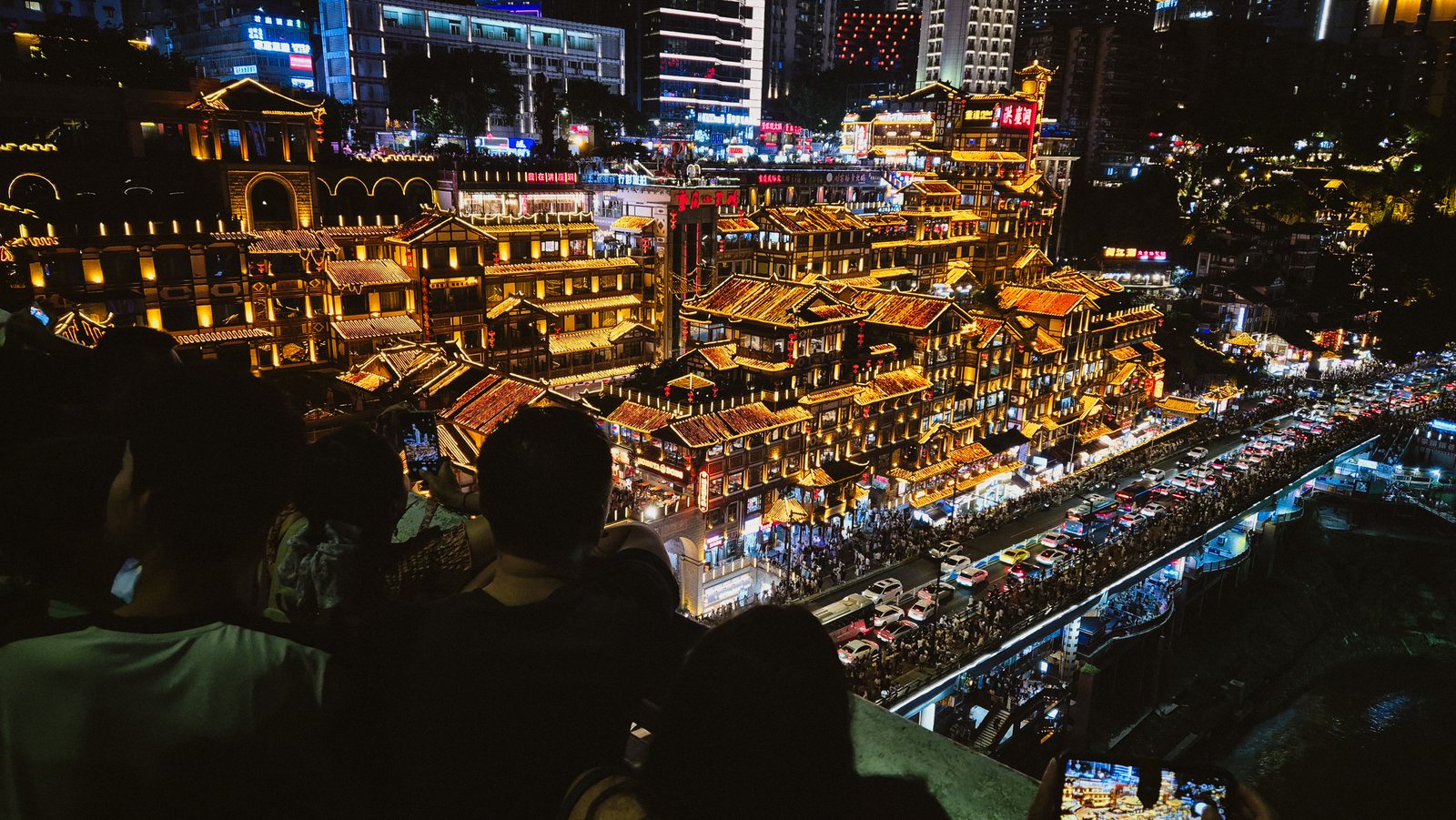 Hongyadong lit up gold at night seen from a viewing platform, with the silhouetted heads of a crowd filling the foreground